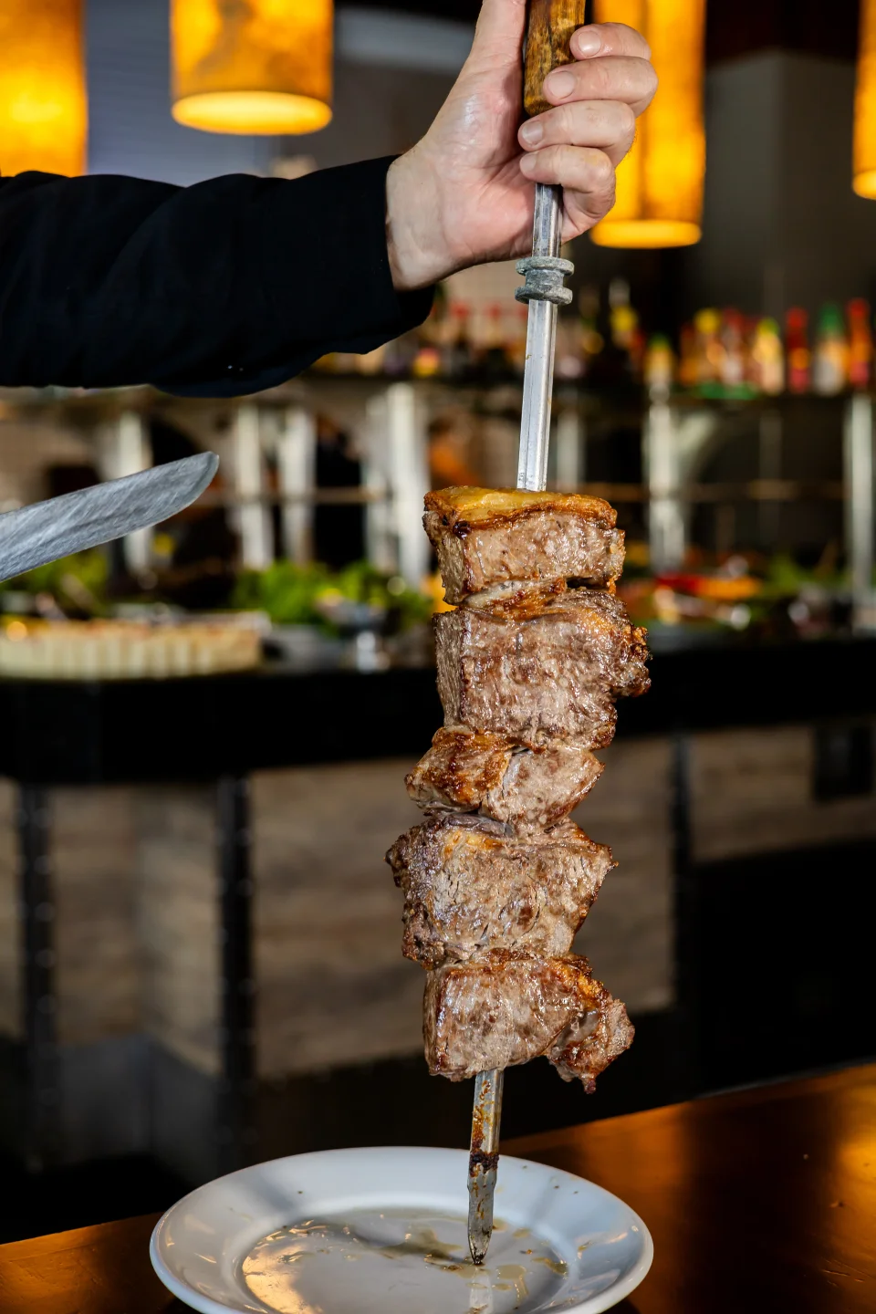 Passador servindo carne no espeto à mesa, com o buffet ao fundo